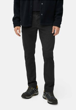Zwarte slim-fit broek, gemaakt van duurzaam materiaal. Draagt een donker topje en stevige schoenen met gele accenten.