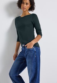 Donkergroene langemouwen top met een bootnek, gecombineerd met lichtblauwe straight-leg jeans met een ritsaccent en voorkeurszakken.