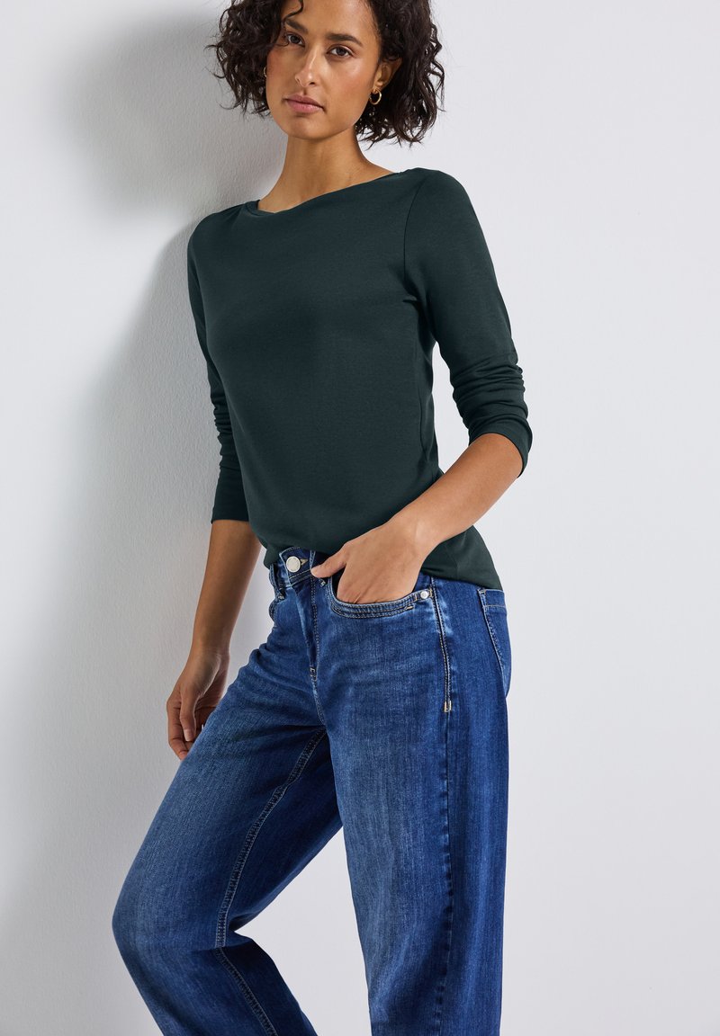 Donkergroene langemouwen top met een bootnek, gecombineerd met lichtblauwe straight-leg jeans met een ritsaccent en voorkeurszakken.