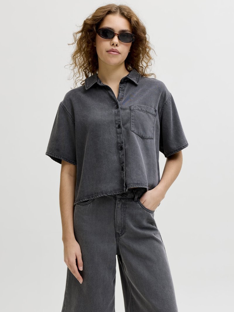 Vrouw met krullend haar, zwarte zonnebril, een loszittend donkergrijs overhemd met korte mouwen en bijpassende hoge taille broek, hand in de zak.