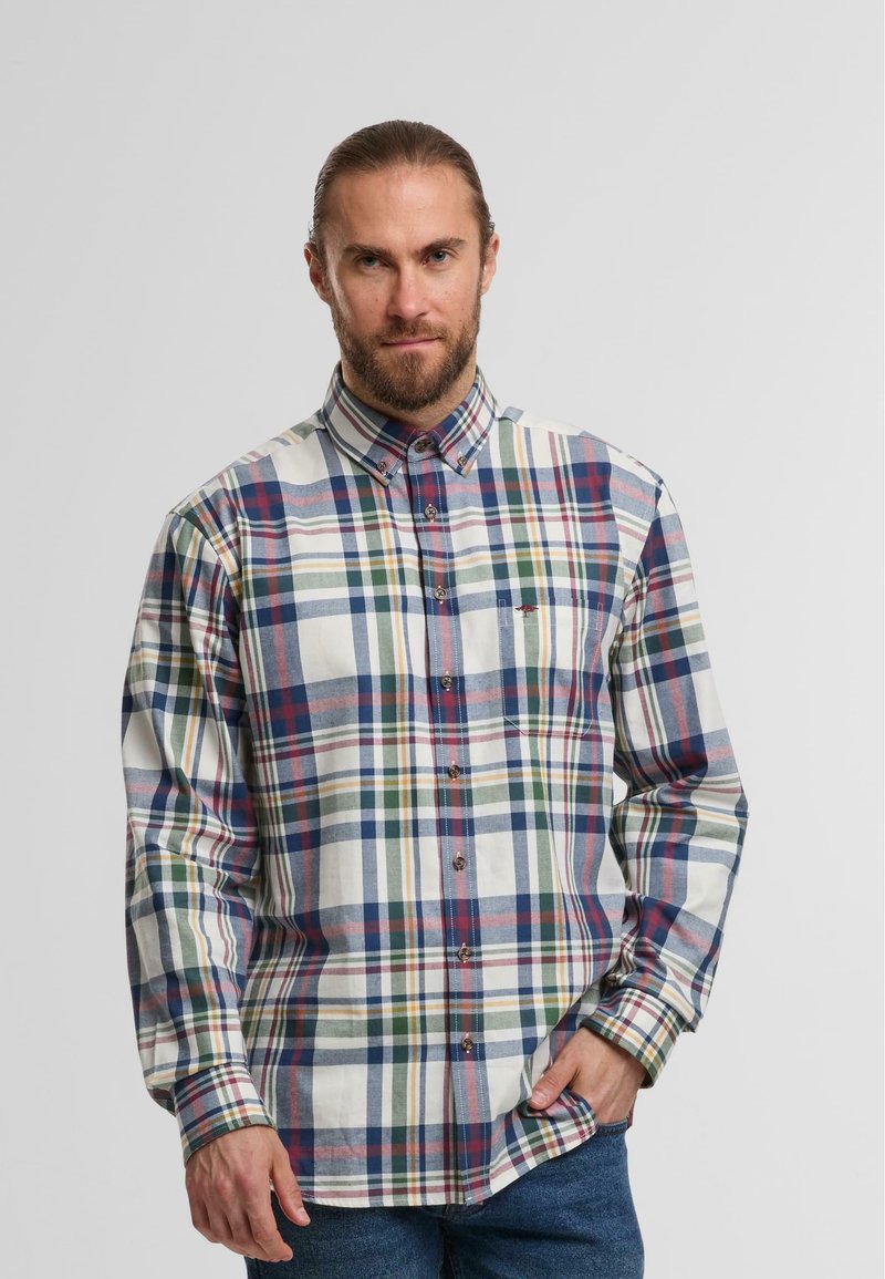 Mann mit Bart, der ein mehrfarbiges karierter Langarmhemd und eine blaue Jeans trägt, steht mit einer Hand in der Tasche.