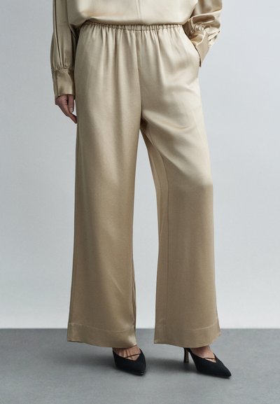 Femme portant un pantalon beige en satin à jambes larges avec taille élastique et des escarpins noirs pointus, debout sur un sol gris devant un fond uni.