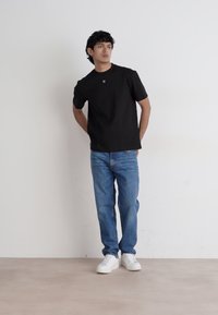 Svart t-skjorte med kort erm og et diskret logo, sammen med lyseblå denimjeans og hvite joggesko. Plassert mot en ensfarget bakgrunn.