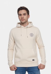 Beżowy hoodie wykonany z miękkiej tkaniny, z kieszenią typu kangur, kapturem ze sznurkiem oraz naszywką z logo na klatce piersiowej. Klasyczny fason z długimi rękawami.