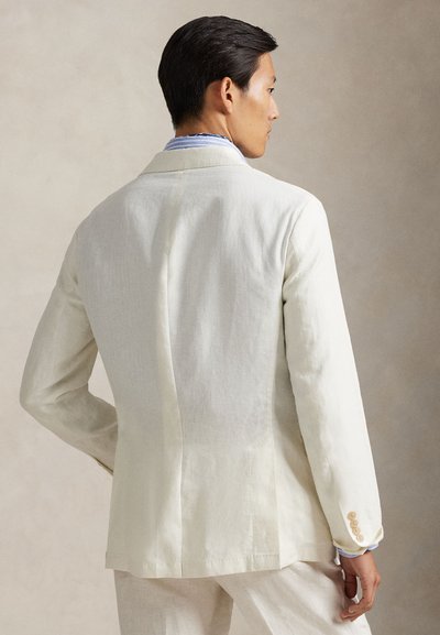 Kreminės spalvos lininis švarkas su lengvai pritaikyta forma, vienu kirpimu ir smėlio spalvos sagomis. Dėvimas ant šviesiai mėlynos, dryžuotos marškinių su apykakle.