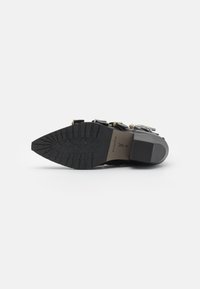 Bota negra hasta el tobillo con suela texturizada, con acentos de hardware dorado y un diseño de tacón grueso. Marca visible en la plantilla.