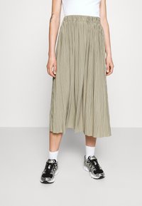Kvinde iført en beige plisseret midi-nederdel, hvide sokker, sorte og sølv sneakers samt en hvid ærmeløs top, stående på et lys gråt gulv.
