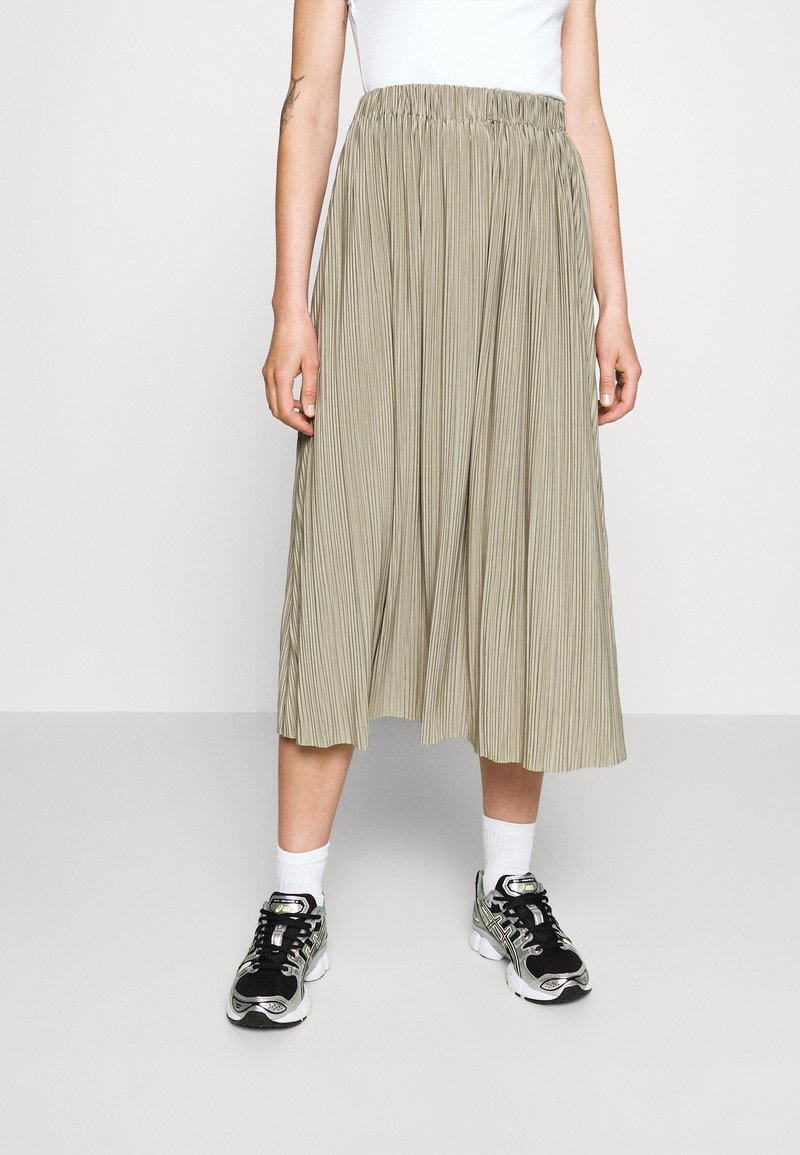 Kvinde iført en beige plisseret midi-nederdel, hvide sokker, sorte og sølv sneakers samt en hvid ærmeløs top, stående på et lys gråt gulv.