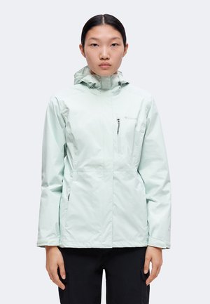 Jeune personne aux cheveux noirs et raides portant une veste de pluie à capuche vert clair avec des poches zippées et un pantalon noir.