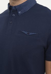Homme portant un polo bleu marine avec un col à motifs et un rabat de poche poitrine à boutons assorti à motifs.