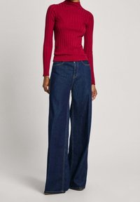 Roter gerippter Rollkragenpullover mit figurbetontem Design, kombiniert mit weit geschnittenen blauen Denim-Jeans mit sichtbaren Nähten und Seitentaschen.