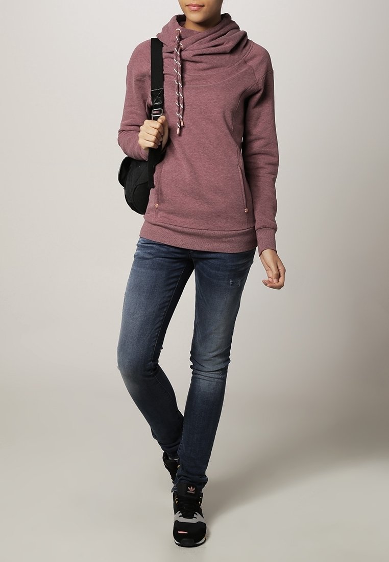 Personne portant un sweat à capuche mauve, un jean bleu, des baskets noires et blanches, et portant un sac à bandoulière noir.