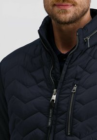 Donker marineblauwe gewatteerde jas met een staande kraag, diagonale chevronstiksels en zilveren ritsen. Voorzien van een gemerkte ritsenpulk en zakdetails.