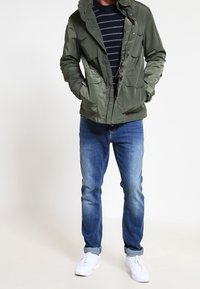 Tmavě zelená, lehká bunda s kapucí a několika kapsami, která je zkombinována se středně modrými džíny a bílými teniskami.