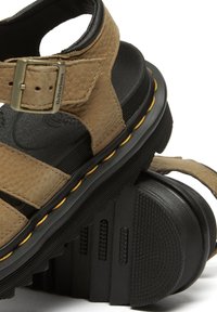 Sandalo in pelle marrone con fibbia regolabile, suola in gomma nera, cinturini testurizzati e dettagli di cucitura gialla lungo i bordi.