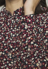 Květinová košile na knoflíky z tmavé látky s červenými, bílými a růžovými květy. Límec je zakulacený s nařasením na ramenech.