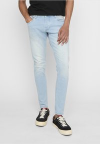 Homme portant un jean slim bleu clair avec des marques d'usure subtiles et des baskets noires à lacets blancs et semelles épaisses, debout, une main dans la poche.