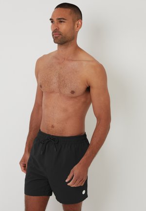 Mężczyzna o krótkich włosach, ubrany w czarne kąpielówki z sznurkiem, stoi na tle jednolitego jasnego tła, spoglądając lekko w prawo.