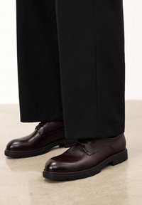 Zapatos de cuero burdeos con una textura suave, punta redondeada y suelas planas negras. Combinados con pantalones negros de pierna ancha.