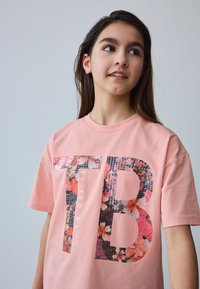 Lyserød bomulds-T-shirt med stort blomsterdesign i bogstaverne "TB" med metallic accenter og rund halsudskæring.
