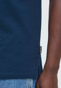 Oseba z roko poleg roba mornarsko modre majice s etiketo "BLEND", v kombinaciji z lahkimi svetlo modrimi trapericami.