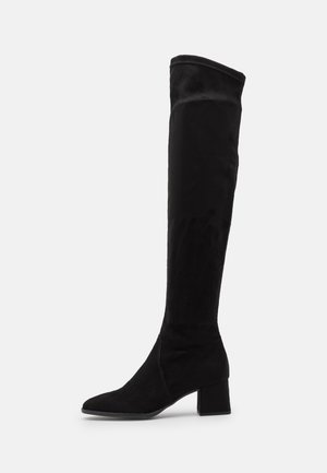 Botte en daim noire jusqu'au genou avec talon bloc et bout pointu, vue de profil sur un fond clair uni.