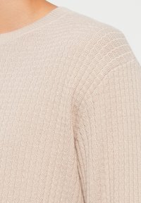 Suéter de punto beige texturizado con escote redondo y un sutil patrón ondulado. Tejido suave con una apariencia ligera, destacando el detalle de la trama.