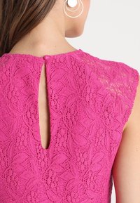 Rosa spetsklänning med en nyckelhålsrygg, med blommönster och en knäppning vid halsen. Mjuk textur och korta ärmar.