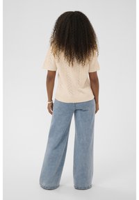 Ženska s kodraste lase, oblečena v bež pleteno majico s kratkimi rokavi in svetlo modre široke kavbojke, stoji obrnjena stran od kamere na svetli podlagi.