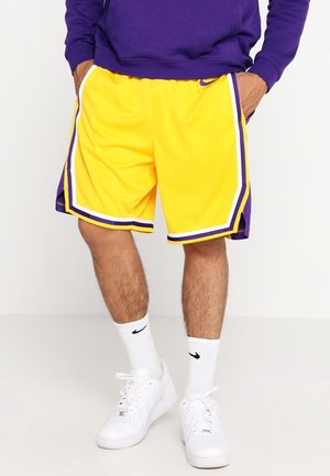 Uomo che indossa pantaloncini da basket gialli e viola, calzini bianchi Nike e sneakers bianche Nike, in piedi con le mani nelle tasche.