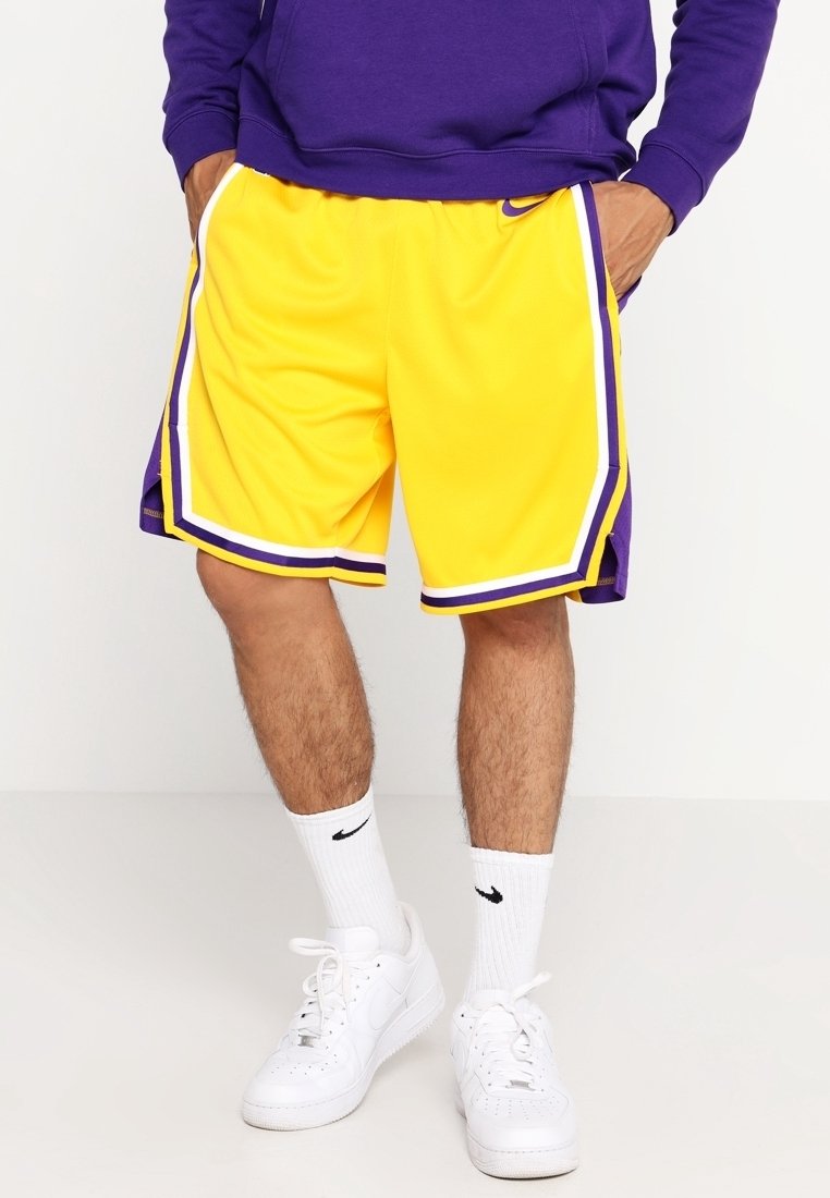 Man wearing yellow and purple basketball shorts, white Nike socks, and white Nike sneakers standing with hands in pockets.