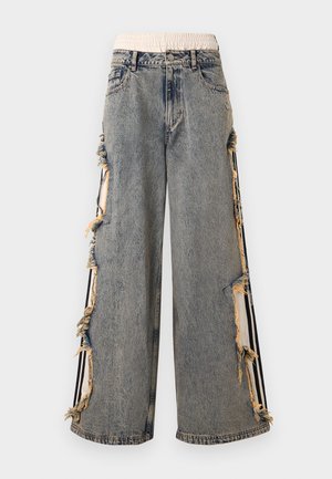Pantalon large en denim gris avec une taille élastique beige, grandes fentes latérales effilochées et accents à rayures noires sur les jambes extérieures.