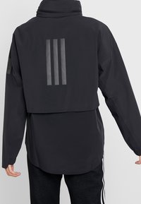 Personne portant une veste noire avec trois bandes réfléchissantes verticales sur le haut du dos et un pantalon noir avec des bandes blanches sur les côtés.