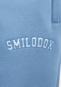 Jasnoniebieska bluza z haftowanym napisem "SMILODOX" w nieco ciemniejszym odcieniu niebieskiego. Materiał ma miękkie, teksturowane wykończenie.