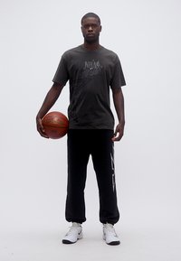 Άνδρας όρθιος που κρατά μια μπάλα μπάσκετ, φορούσε μαύρο t-shirt NBA All-Star, μαύρο παντελόνι φόρμας και λευκά αθλητικά παπούτσια σε απλό φόντο.