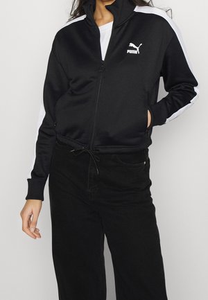 Personne portant une veste zippée noire Puma avec des bandes blanches sur les manches et un pantalon noir, debout avec une main dans la poche.