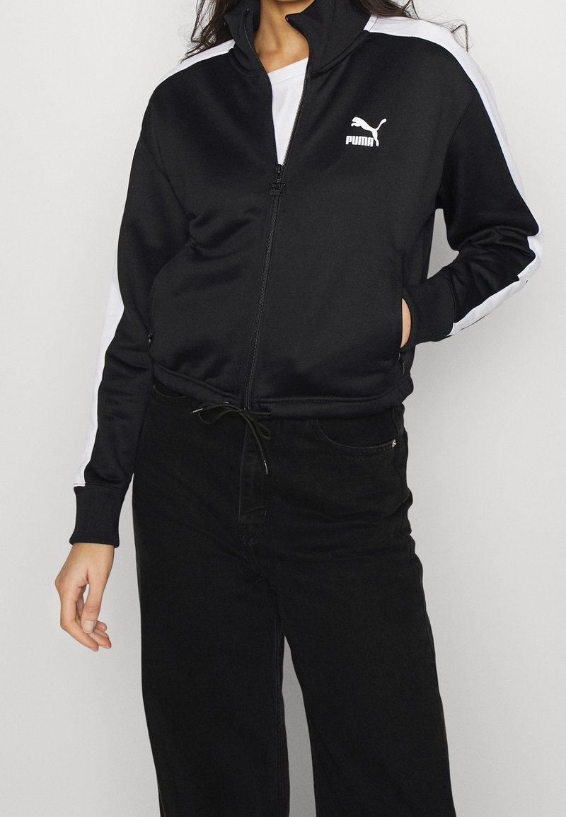 Personne portant une veste zippée noire Puma avec des bandes blanches sur les manches et un pantalon noir, debout avec une main dans la poche.