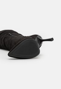 Botte noire en suède à hauteur de genou, avec un bout pointu, une semelle en caoutchouc texturée, un design épuré et un discret détail de fermeture éclair latérale.