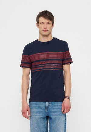 Homme portant un T-shirt bleu marine avec des rayures rouges horizontales sur la poitrine et les manches, un jean bleu clair et une montre-bracelet, debout devant un fond blanc.