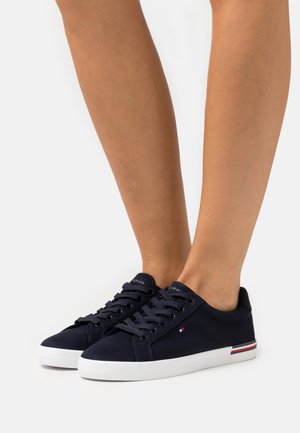 Des jambes portant des baskets en toile bleu marine avec des semelles blanches et des accents rayés rouge-blanc-bleu, se tenant sur un fond blanc uni.