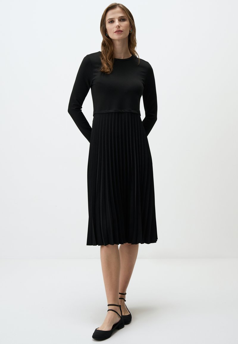 Robe noire avec des manches longues, jupe plissée et corsage ajusté. Fabriquée en tissu lisse, avec un col rond et une coupe longueur cheville.
