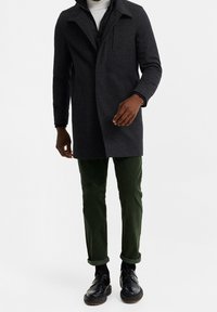 Tmavě šedý kabát z vlněné směsi s límcem, předním zipem a bočními kapsami, doplněný zelenými manšestrovými kalhotami a černými šněrovacími botami.