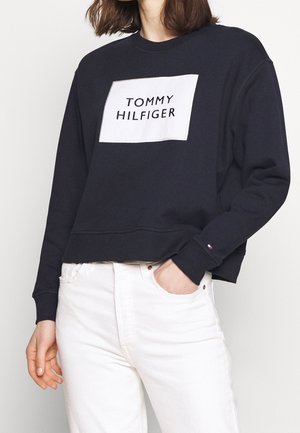 Personne portant un sweatshirt noir Tommy Hilfiger avec un patch logo blanc, assorti à un jean blanc, une main dans la poche.