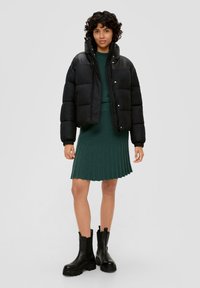 Schwarze Pufferjacke mit hohem Kragen, getragen über einem dunkelgrünen, gerippten Strickkleid. Schwarze Ankle Boots mit auffälligen Sohlen. Einfarbig grauer Hintergrund.