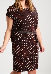 Brun klänning med abstrakta tan-mönster, korta ärmar och en avslappnad passform, åtsittande i midjan med ett flätat bälte. Slät tygkänsla.