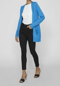 Blauer langer Cardigan über einem weißen Oberteil, kombiniert mit schwarzen Skinny-Jeans und schwarzen offenen High Heels. Der Cardigan hat Taschen und einen lässigen Schnitt.