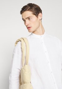 Camisa blanca de lino con un cuello alto, que presenta detalles de botones. Un bolso acolchado beige cuelga del hombro, añadiendo una textura contrastante.
