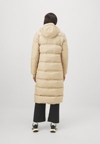 Cappotto imbottito beige con sezioni a righe orizzontali, cappuccio e design a lunghezza intera. Abbinato a pantaloni neri a gamba larga e scarpe sportive.