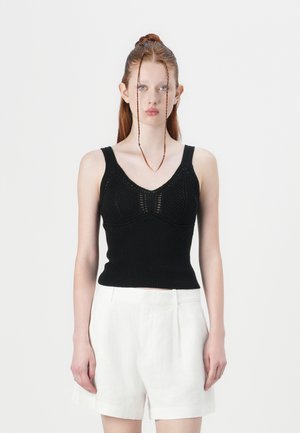 Schwarzes, gestricktes Tanktop mit taillierter Passform, V-Ausschnitt und strukturierten Mustern. Kombiniert mit weißen Leinen-Shorts.