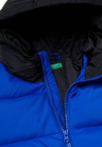 Modrá péřová bunda s černou podšívkou, s plnou zipovou přední částí a korálkovými detaily, a zelenou značkovou visačkou. Hladký povrch.